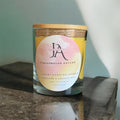 A clear glass jar with a wooden lid labeled 'Josie's Aroma Gingerbread Dreams Luxury Scented Candle', placed on a marble surface with a shadow falling over it.