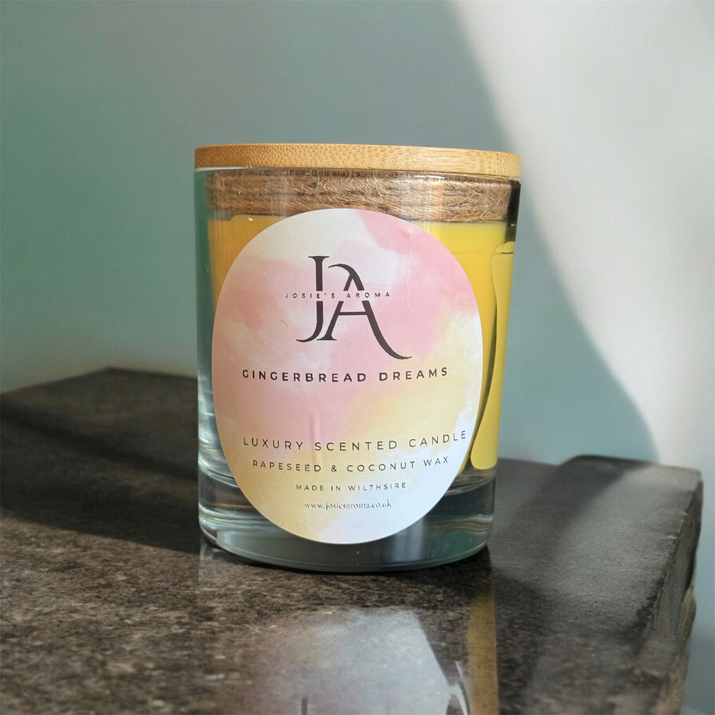 A clear glass jar with a wooden lid labeled 'Josie's Aroma Gingerbread Dreams Luxury Scented Candle', placed on a marble surface with a shadow falling over it.