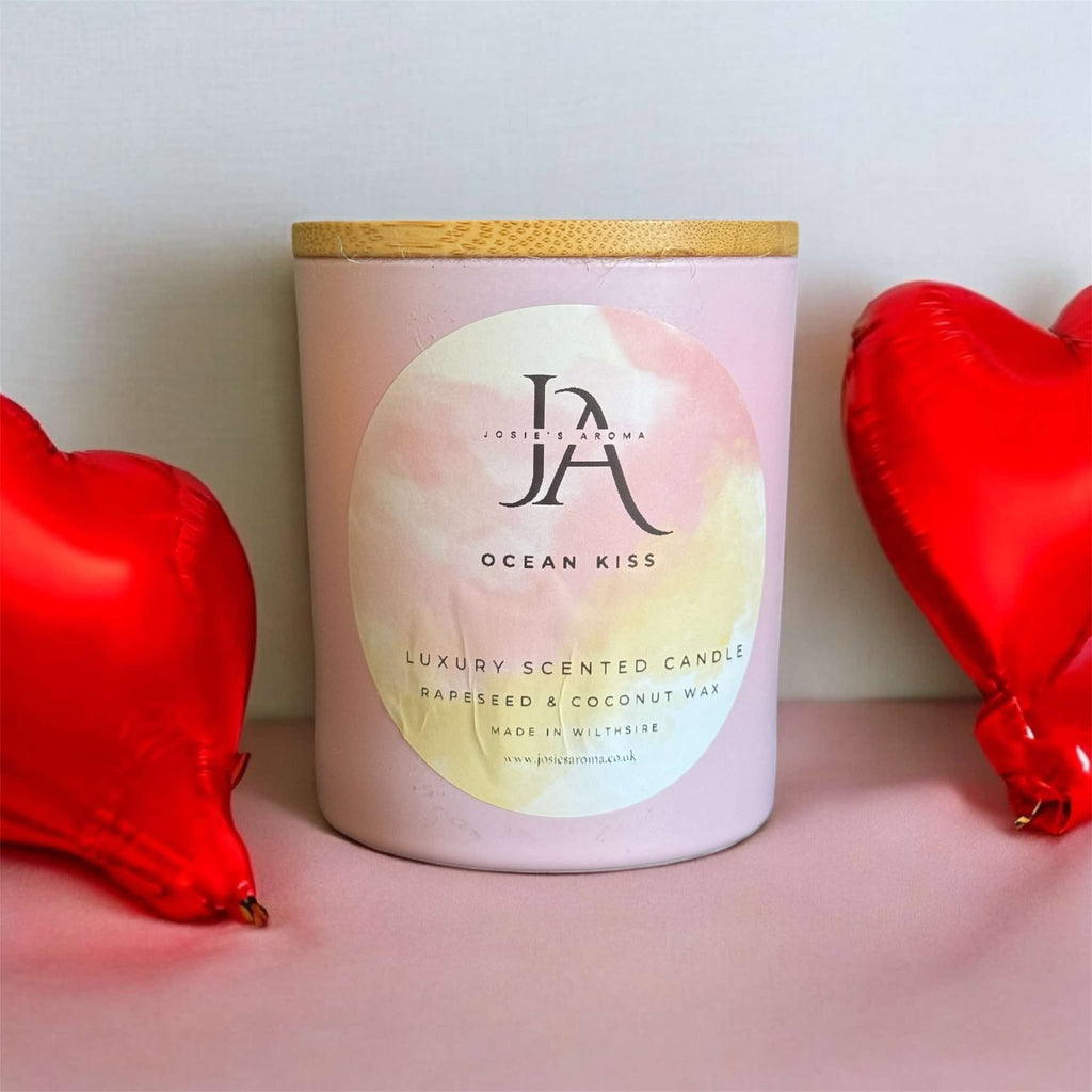A candle with a pink and beige label called 'Ocean Kiss' placed on a surface with red heart-shaped balloons and decorative hearts in the background.