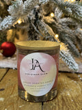 A candle with a label reading 'Josie's Christmas Glow' placed on a wooden surface with a blurred Christmas tree and lights in the background.