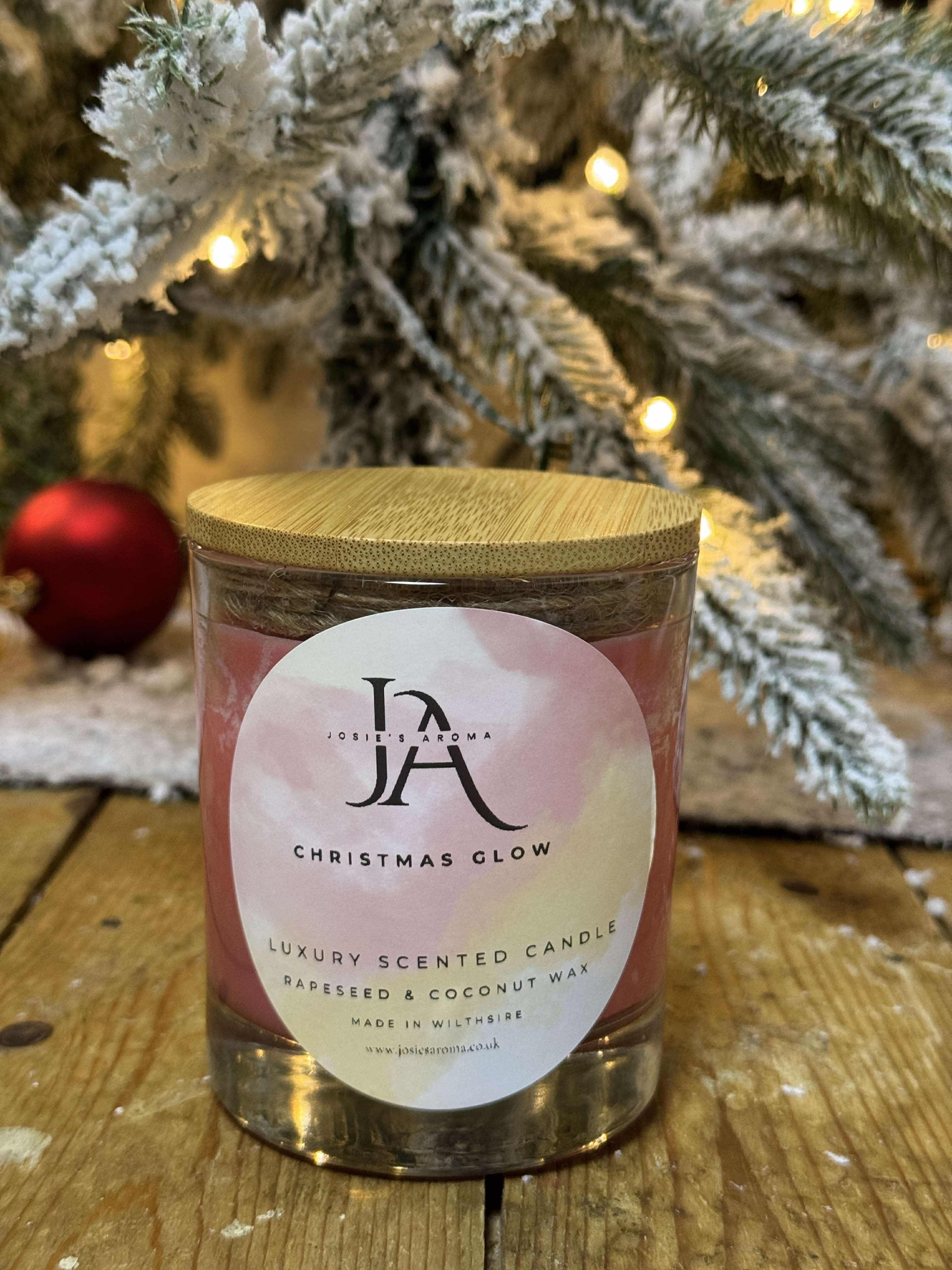 A candle with a label reading 'Josie's Christmas Glow' placed on a wooden surface with a blurred Christmas tree and lights in the background.