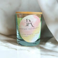 A clear glass candle with a wooden lid, labeled 'Josie's Aroma Christmas Tree Serenity', placed on a marble surface with a white and pink floral fabric in the background.