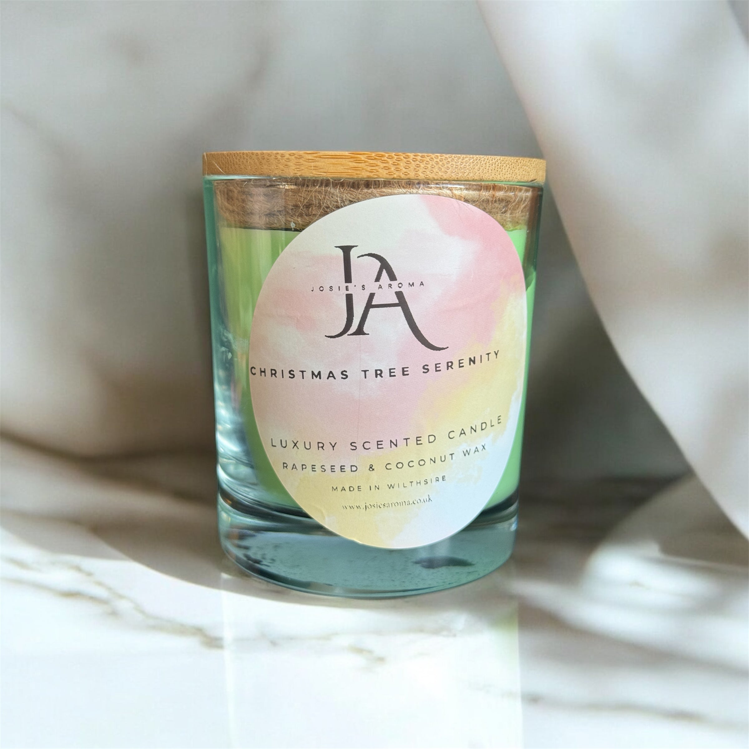 A clear glass candle with a wooden lid, labeled 'Josie's Aroma Christmas Tree Serenity', placed on a marble surface with a white and pink floral fabric in the background.