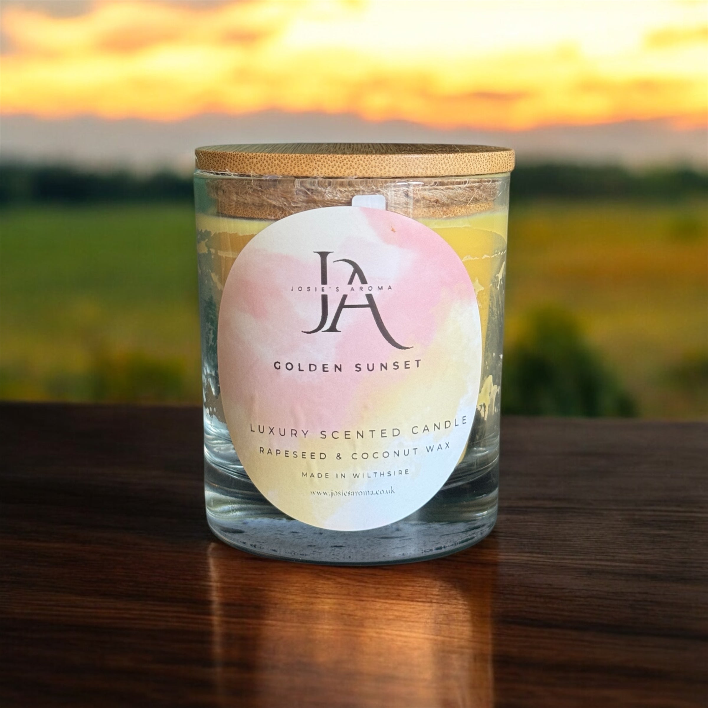 A scented candle with a label reading 'Golden Sunset' in a clear glass jar with a beige lid, set against a rural backdrop.