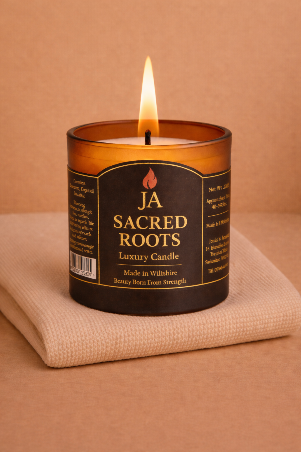 Lit JA Sacred Roots luxury amber candle on folded beige fabric with warm brown background, calming and grounding aroma.