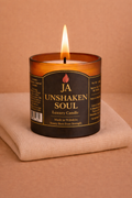 Burning luxury candle labeled Unshaken Soul with amber glass on folded beige fabric background