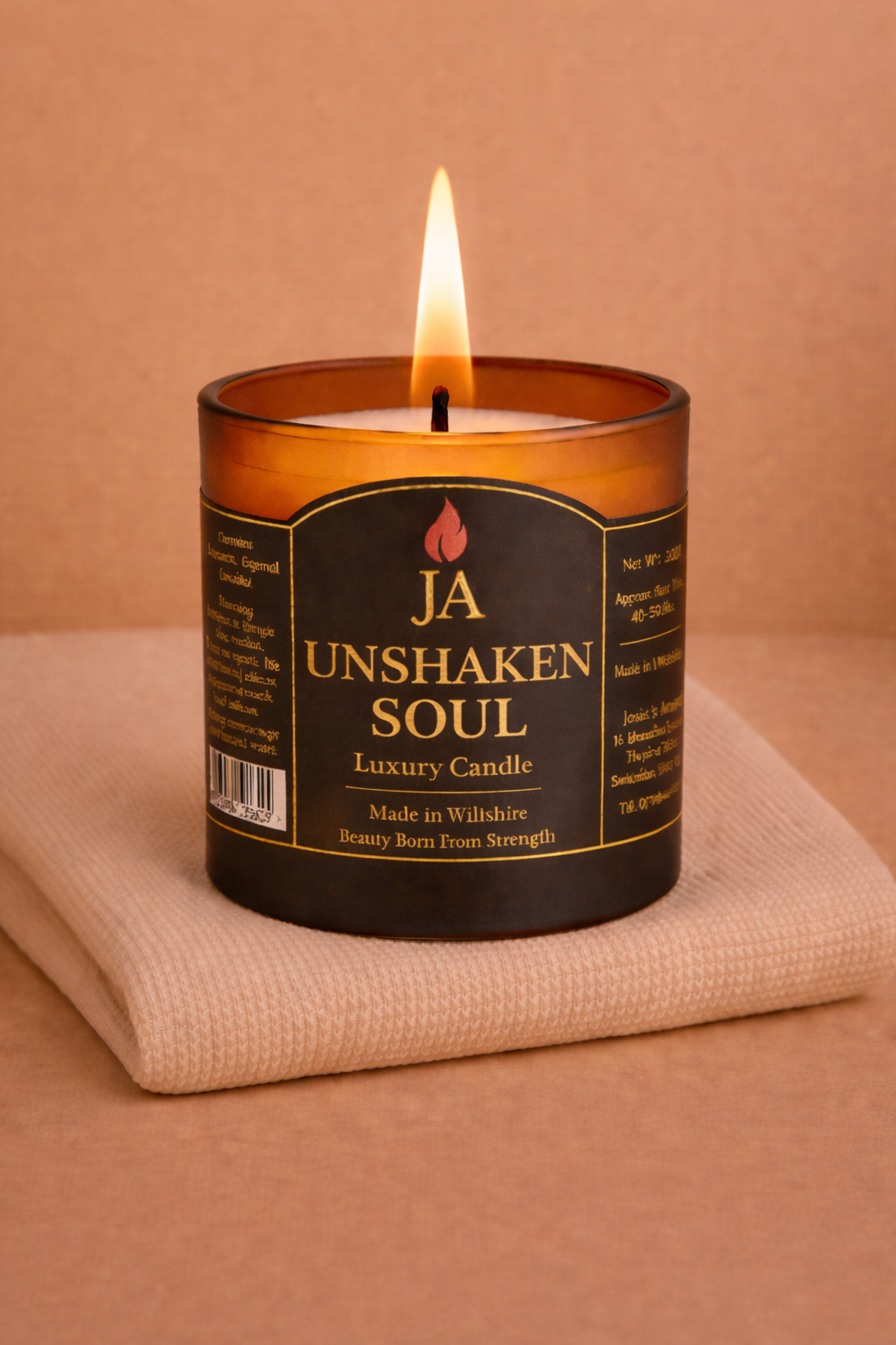 Burning luxury candle labeled Unshaken Soul with amber glass on folded beige fabric background