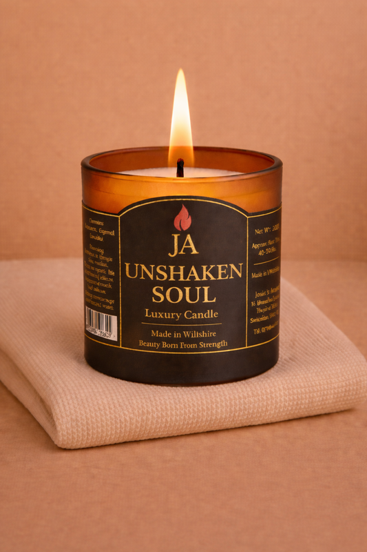 Burning luxury candle labeled Unshaken Soul with amber glass on folded beige fabric background