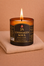 Burning luxury candle labeled Unshaken Soul with amber glass on folded beige fabric background